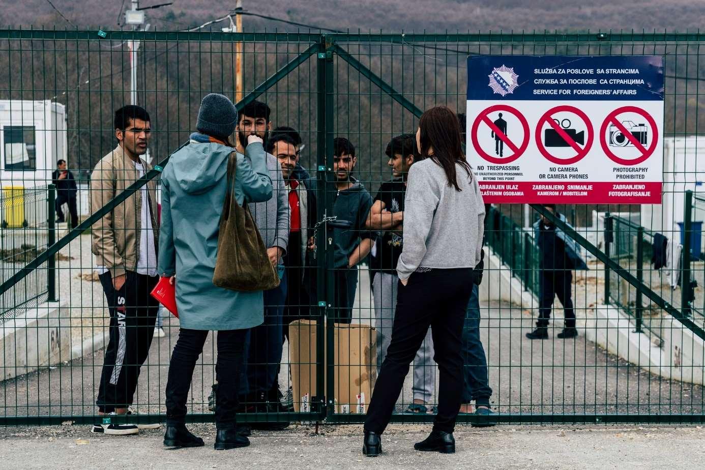 Mensche Sprechen mit Geflüchteten die hinter dem versperrten grünen Metallzaun stehen. Am Zaun ist ein Schild montiert, dass man keine Fotos machen darf.