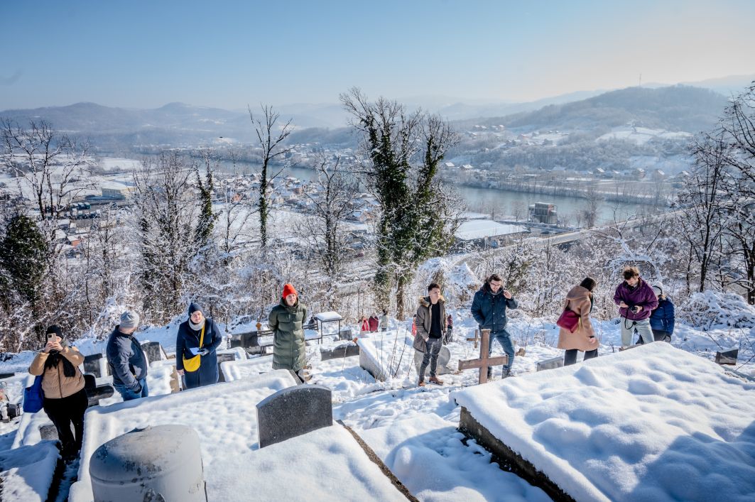 Menschen stehen in einem Halbkreis um tief verschneite Gräber in steilem Gelände. Blauer Himmel. Dahinter eine tief verschneite gebirgige Landschaft mit einem breiten Fluss.